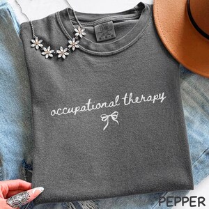 May include: A charcoal gray t-shirt with the words "occupational therapy" in white script, with a bow graphic underneath. A silver necklace with flower-shaped pendants and a brown hat are also visible.