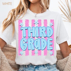 May include: White t-shirt with a pink and blue design. The design features the words "THIRD GRADE" in large, light blue, bubble letters, set against a pink striped background. Small blue stars are scattered around the text.