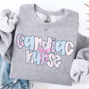 K&ouml;nnte beinhalten: Ein graues Sweatshirt mit dem Schriftzug "Cardiac Nurse" in einem farbenfrohen Muster. Die Buchstaben sind mit blauen Streifen, rosa Vichy-Karo und floralen Mustern gef&uuml;llt. Eine rosa Schleife ist auf dem Wort "Nurse" zentriert. Eine silberne Halskette ist ebenfalls sichtbar.