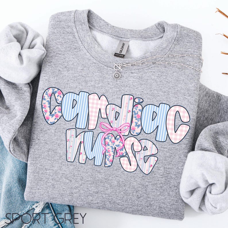 K&ouml;nnte beinhalten: Ein graues Sweatshirt mit dem Schriftzug "Cardiac Nurse" in einem farbenfrohen Muster. Die Buchstaben sind mit blauen Streifen, rosa Vichy-Karo und floralen Mustern gef&uuml;llt. Eine rosa Schleife ist auf dem Wort "Nurse" zentriert. Eine silberne Halskette ist ebenfalls sichtbar.