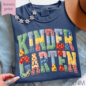 May include: A navy blue crew neck t-shirt with the word "KINDERGARTEN" in colorful patchwork letters. The shirt has a relaxed fit and is made of a soft material. A floral necklace and a brown hat are also visible.