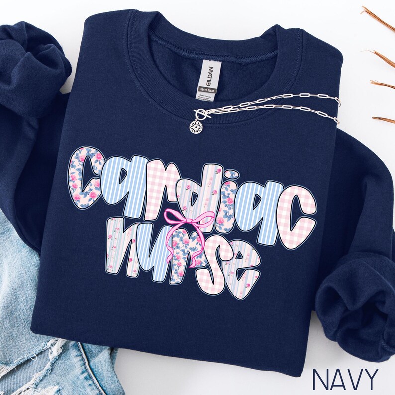 K&ouml;nnte beinhalten: Marineblaues Sweatshirt mit dem Schriftzug "Cardiac Nurse" in einer verspielten Schriftart. Die Buchstaben sind mit floralen und Vichy-Mustern in Rosa und Blau verziert. Eine rosa Schleife verziert das Design. Eine silberne Halskette liegt &uuml;ber dem Sweatshirt.