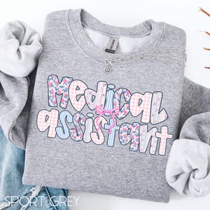 May include: A grey sweatshirt featuring the text "Medical Assistant" in a bubble font. The letters are decorated with floral, gingham, and striped patterns in pink and blue. A pink bow is between the words. A silver necklace is on the sweatshirt.