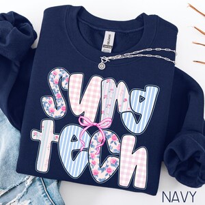 May include: Navy blue sweatshirt with the text "SUNY TECH" in a playful font. The letters feature floral, gingham, and striped patterns in pink and blue. A silver necklace with a pendant is draped across the sweatshirt.