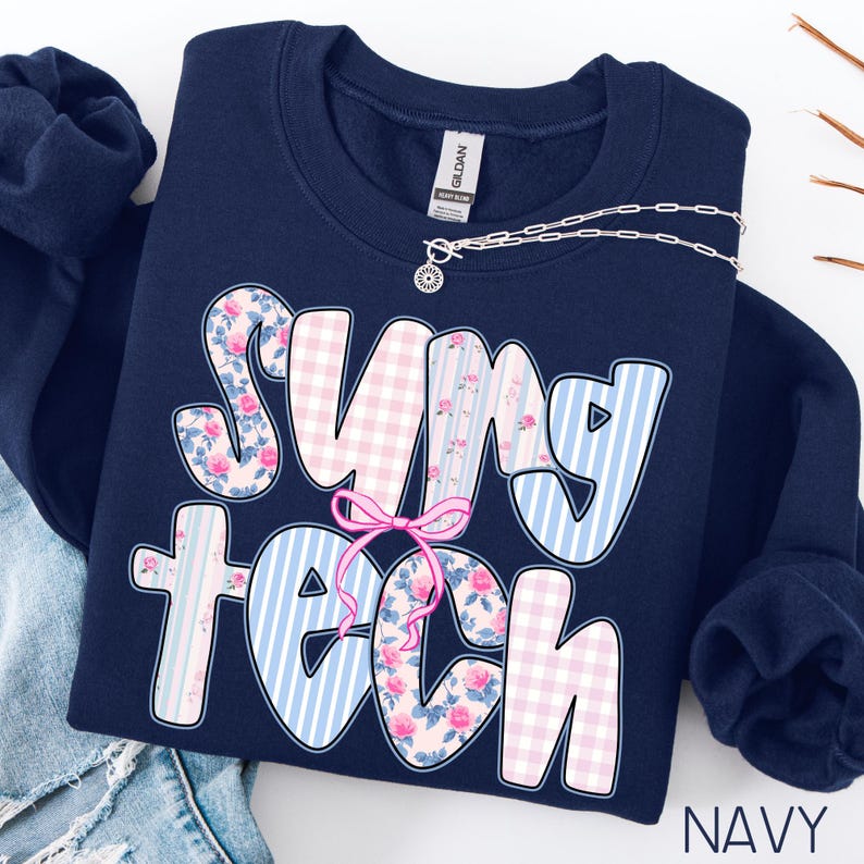 May include: Navy blue sweatshirt with the text "SUNY TECH" in a playful font. The letters feature floral, gingham, and striped patterns in pink and blue. A silver necklace with a pendant is draped across the sweatshirt.