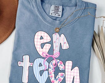 May include: A blue jean colored t-shirt with the words "EM TECH" in a playful font. The letters are decorated with pink gingham, floral patterns, and blue stripes. A pink bow is between the letters "EM" and "TECH". A gold necklace is draped across the shirt.