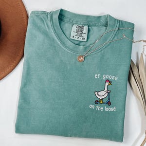 Peut inclure: T-shirt Comfort Colors vert d'eau avec le texte "er goose on the loose" et une oie brodée sur un scooter. Un collier argenté avec un pendentif est sur le t-shirt.