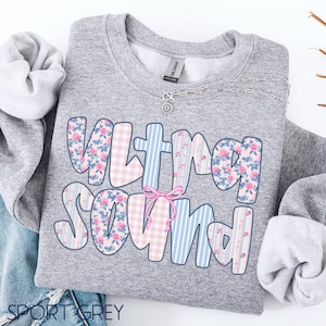 May include: A grey sweatshirt featuring the words "ULTRA SOUND" in a playful font. The letters are decorated with pink floral, blue striped, and pink gingham patterns. A pink bow is between the letters. A silver necklace is on the sweatshirt.