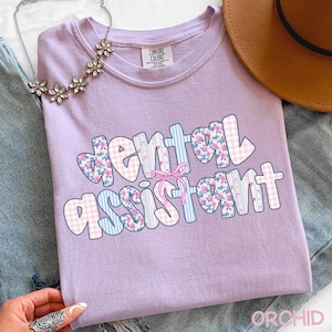 Pode incluir: Uma t-shirt lil&aacute;s com a frase "Dental Assistant" numa fonte colorida e divertida. As letras s&atilde;o decoradas com padr&otilde;es florais, xadrez e listrados. Um la&ccedil;o rosa acentua o design.
