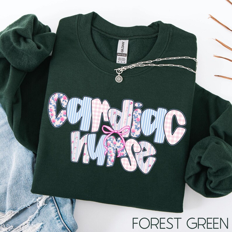 K&ouml;nnte beinhalten: Ein dunkelgr&uuml;nes Sweatshirt mit den Worten "Cardiac Nurse" in einem floralen und gestreiften Design. Eine silberne Halskette mit einem kleinen Anh&auml;nger befindet sich auf dem Sweatshirt. Die Worte "Forest Green" stehen am unteren Rand des Bildes.