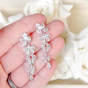 May include: A pair of silver-toned, dangling earrings with a floral design. Each earring features multiple clear, sparkling, marquise-cut and round-cut crystals arranged in a cascading pattern. The earrings are held against a soft, blurred background.