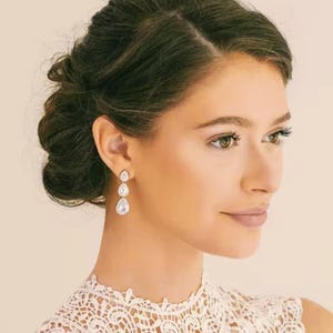 Peut inclure: Une femme avec les cheveux tirés en arrière en chignon porte un haut en dentelle blanche et des boucles d'oreilles en cristal pendantes. Les boucles d'oreilles sont en forme de goutte et ont une teinte argentée.