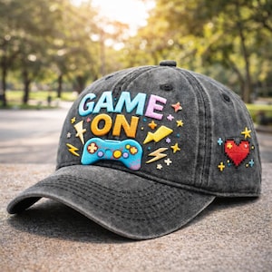 May include: A grey denim baseball cap with the words "GAME ON" in colourful, raised lettering. The cap features a blue and teal game controller, lightning bolts, stars, and a pixelated red heart.
