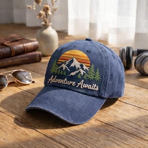 Może przedstawiać: Granatowa czapka bejsbolowa z haftowanym wzorem gór i zachodu słońca. Czapka ma napis "Adventure Awaits" w białym kolorze. Kapelusz leży na drewnianej powierzchni.