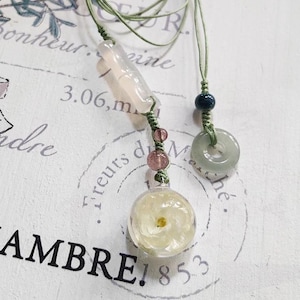 May include: A delicate necklace with a green cord, featuring a light pink and white rectangular pendant, a round pendant with a white flower inside, and a green jade ring pendant. The necklace also includes small pink and blue beads.