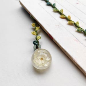 May include: A white flower encased in a clear glass sphere, attached to a green and yellow crocheted vine bookmark.