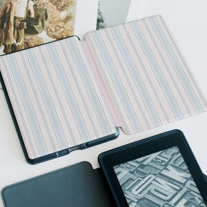 May include: An open e-reader case with a pink and blue striped pattern. The case is open, revealing the interior and the e-reader screen. The e-reader is black, and the case has a dark blue border.