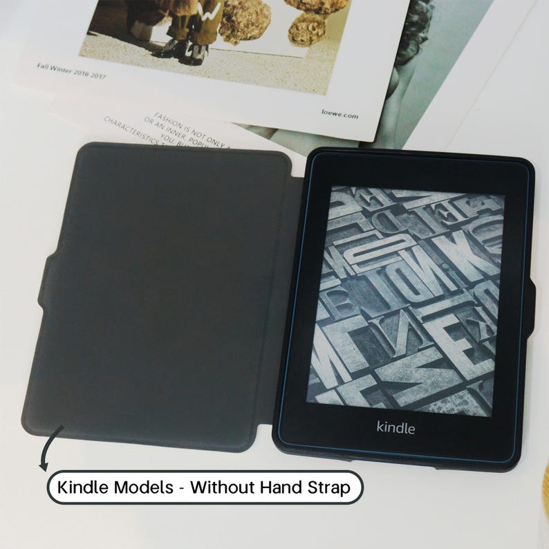 May include: A black Kindle e-reader with a dark gray cover, open to display a screen of gray letterforms. The Kindle is on a white surface with a magazine in the background. Text on the image reads "Kindle Models - Without Hand Strap."