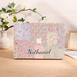 May include: A floral patterned laptop case with a patchwork design in shades of pink, blue, and yellow. The name "Nathaniel" is printed in a cursive font on the bottom. The Apple logo is visible.