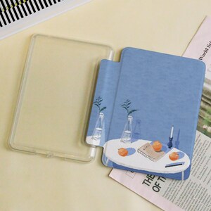 May include: A clear, protective tablet case next to a blue tablet cover with a painted illustration. The illustration depicts a white table with oranges, a candle, and vases with plants. The text "THE OF T" is visible.