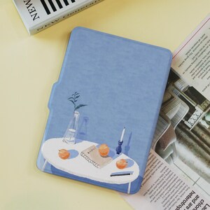 May include: A light blue tablet case featuring a painted illustration of a small white table with oranges, a notebook, a candle, and a vase with a plant. The word "NEW" is visible on a book in the background.
