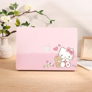 May include: A pink laptop case featuring a Hello Kitty design with a teddy bear, hearts, and candy. The case has a glossy finish and a cutout for the Apple logo. The design is in a cartoon style.