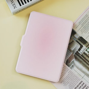 May include: A light pink tablet case with a smooth surface. The case has a rectangular shape with rounded corners and a small cutout on the left side. A book with the word "NEW" is visible in the upper left corner.