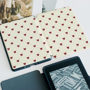 May include: Cream-colored e-reader cover with a repeating pattern of red hearts connected by dotted lines. The cover is open, revealing the screen of the e-reader. The e-reader is black.