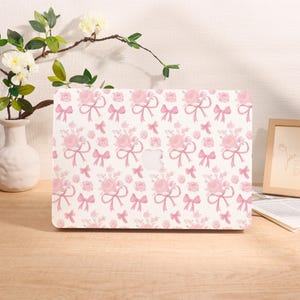 May include: A white laptop case featuring a repeating pattern of pink roses and bows. The design includes bouquets of roses tied with ribbons and scattered individual roses and bows. The case has a cutout for the Apple logo.