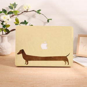 Könnte beinhalten: Hellgelbe Laptop-Hülle mit einer braunen Dackel-Illustration. Die Hülle hat ein weißes Apple-Logo und steht auf einer Holzoberfläche. Eine weiße Vase mit Blumen und ein gerahmtes Bild befinden sich im Hintergrund.