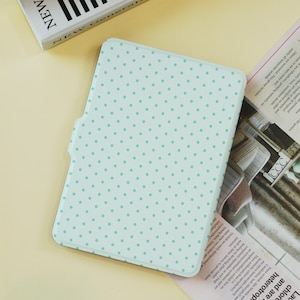 Puede incluir: Una funda de tableta verde claro con un patrón de lunares. La funda tiene un cierre magnético y es de forma rectangular. Los puntos son de un tono de verde más claro. La palabra "NEW" es visible en un libro en el fondo.