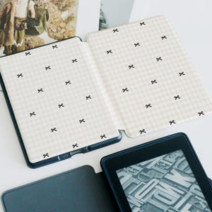 May include: A white and pink gingham patterned e-reader case with black bow accents. The case is open and the e-reader is visible with a black and white text screen.