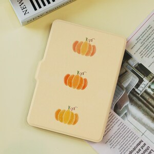 Può includere: Una custodia per tablet beige chiaro con tre illustrazioni di zucche arancioni e gialle. Le zucche hanno steli verdi e sono disposte verticalmente. La custodia è probabilmente per prodotti a tema autunnale.