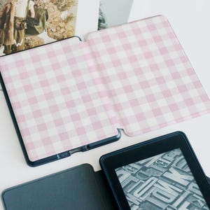 May include: A black e-reader with a pink and white gingham patterned cover, open to display the screen. The cover is open, revealing the screen with a display of letter blocks. The cover is made of a soft material.