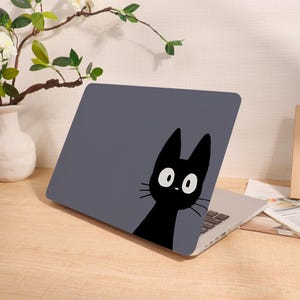 May include: A laptop case with a dark gray background featuring a black cat illustration. The cat has large white eyes and a small nose. The laptop is open on a wooden surface.