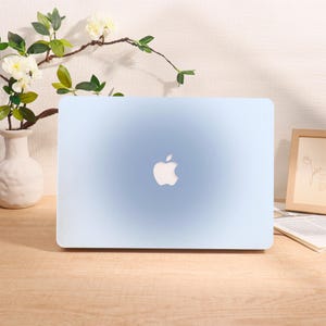 Könnte beinhalten: Ein hellblaues Laptop mit dem Apple-Logo auf dem Deckel. Der Laptop steht auf einer Holzoberfläche, mit einer weißen Vase mit Blumen und einem gerahmten Bild im Hintergrund.