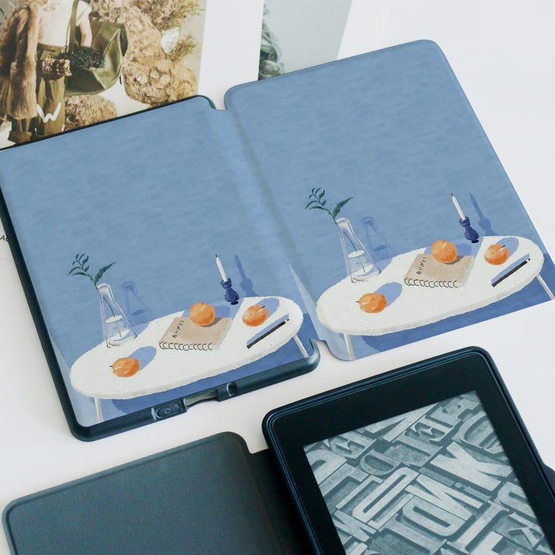 May include: A blue Kindle case with a watercolor illustration of a table with oranges, a candle, and a vase. The case is open, revealing the screen of the Kindle and the illustrated design.