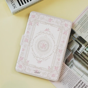 Puede incluir: Una funda para tableta con estampado floral blanco y rosa con cierre magnético. La funda presenta un diseño detallado con un medallón floral central y la palabra "Violet" en escritura. La funda es rectangular y tiene esquinas redondeadas.