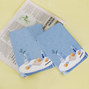 May include: Three light blue notebooks with a watercolor illustration of a table with oranges, a vase, and a pen. The notebooks are on top of a newspaper with the text "SPECIFIC INTRODUCTION".