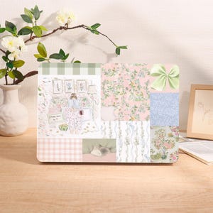 May include: A decorative laptop skin featuring a patchwork design with floral, gingham, and bow patterns. The design includes a watercolor illustration of a person in pajamas reading in a room, with a cat sleeping.