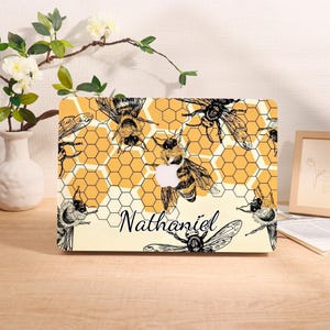 Könnte beinhalten: Eine Laptop-Hülle mit einem Bienen- und Wabenmuster. Die Hülle hat einen gelben Hintergrund mit schwarz-weißen Illustrationen von Bienen und Waben. Der Name "Nathaniel" ist in Kursivschrift auf der Hülle geschrieben.