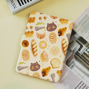 May include: A cream-colored tablet case featuring a repeating pattern of baked goods, cat faces, and paw prints. The case has a rounded shape and a cutout for a camera. The design includes croissants, bread loaves, and other pastries.