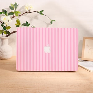 May include: Pink laptop with a vertical striped pattern. The laptop has a white Apple logo in the center. The background includes a white vase with flowers and a framed picture.
