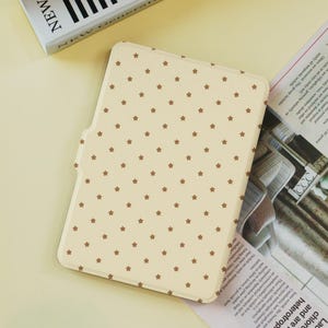 Może przedstawiać: Kremowe etui na tablet z powtarzającym się wzorem małych brązowych gwiazdek. Etui posiada magnetyczne zamknięcie i jest przeznaczone do ochrony tabletu. Etui jest prostokątne z zaokrąglonymi rogami.