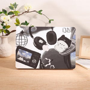 May include: A white laptop case with a black and white illustration featuring a camera, headphones, a globe, and a flip phone. The design includes cartoon-style elements and a light gray background.