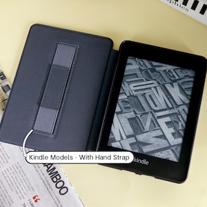May include: Black Kindle e-reader with a gray hand strap, displayed open in a black case. The screen shows a gray and white pattern. The text "Kindle Models - With Hand Strap" is visible.