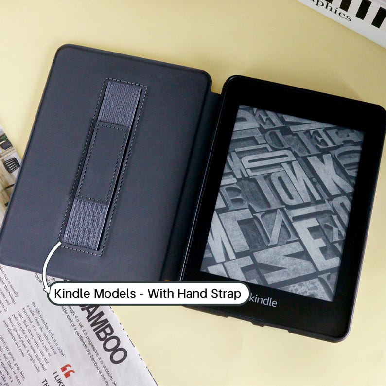 May include: Black Kindle e-reader with a gray hand strap, displayed open in a black case. The screen shows a gray and white pattern. The text "Kindle Models - With Hand Strap" is visible.