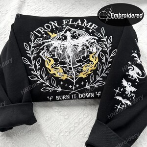 K&ouml;nnte beinhalten: Schwarzer Sweatshirt mit einem wei&szlig;en gestickten Design eines Drachen mit Flammen und dem Text "IRON FLAME" und "BURN IT DOWN". Das Design ist von einem Kranz aus Bl&auml;ttern umgeben. Die &Auml;rmel haben kleinere gestickte Designs von Drachen.
