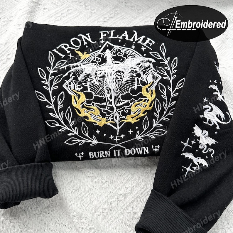 K&ouml;nnte beinhalten: Schwarzer Sweatshirt mit einem wei&szlig;en gestickten Design eines Drachen mit Flammen und dem Text "IRON FLAME" und "BURN IT DOWN". Das Design ist von einem Kranz aus Bl&auml;ttern umgeben. Die &Auml;rmel haben kleinere gestickte Designs von Drachen.