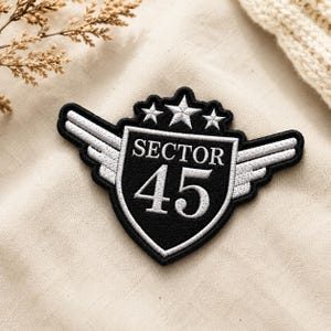 May include: Embroidered patch in black and white, shaped like a shield with wings. The patch reads "SECTOR 45" and has three stars at the top. The wings are white and the background is a neutral tone.